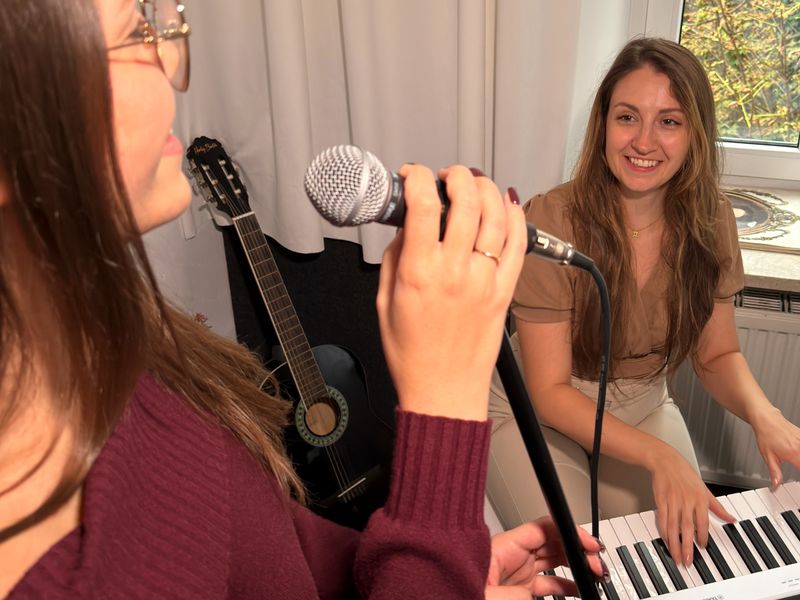 Klaudia Kita uśmiecha się, grając na pianinie podczas lekcji śpiewu; na pierwszym planie uczennica śpiewa do mikrofonu